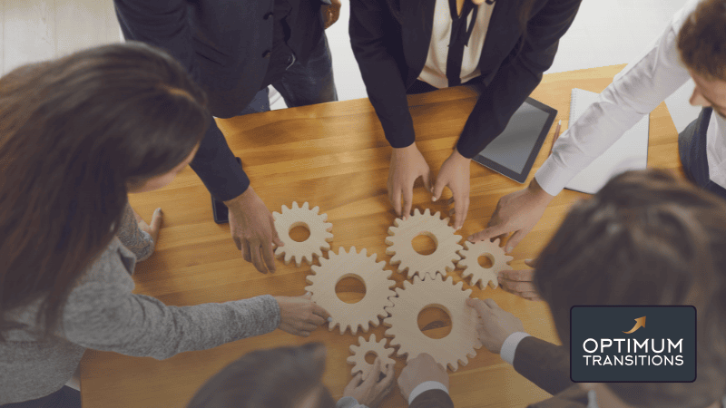 A team of employees stands around a wooden table, fitting together wooden cogs to make a working system